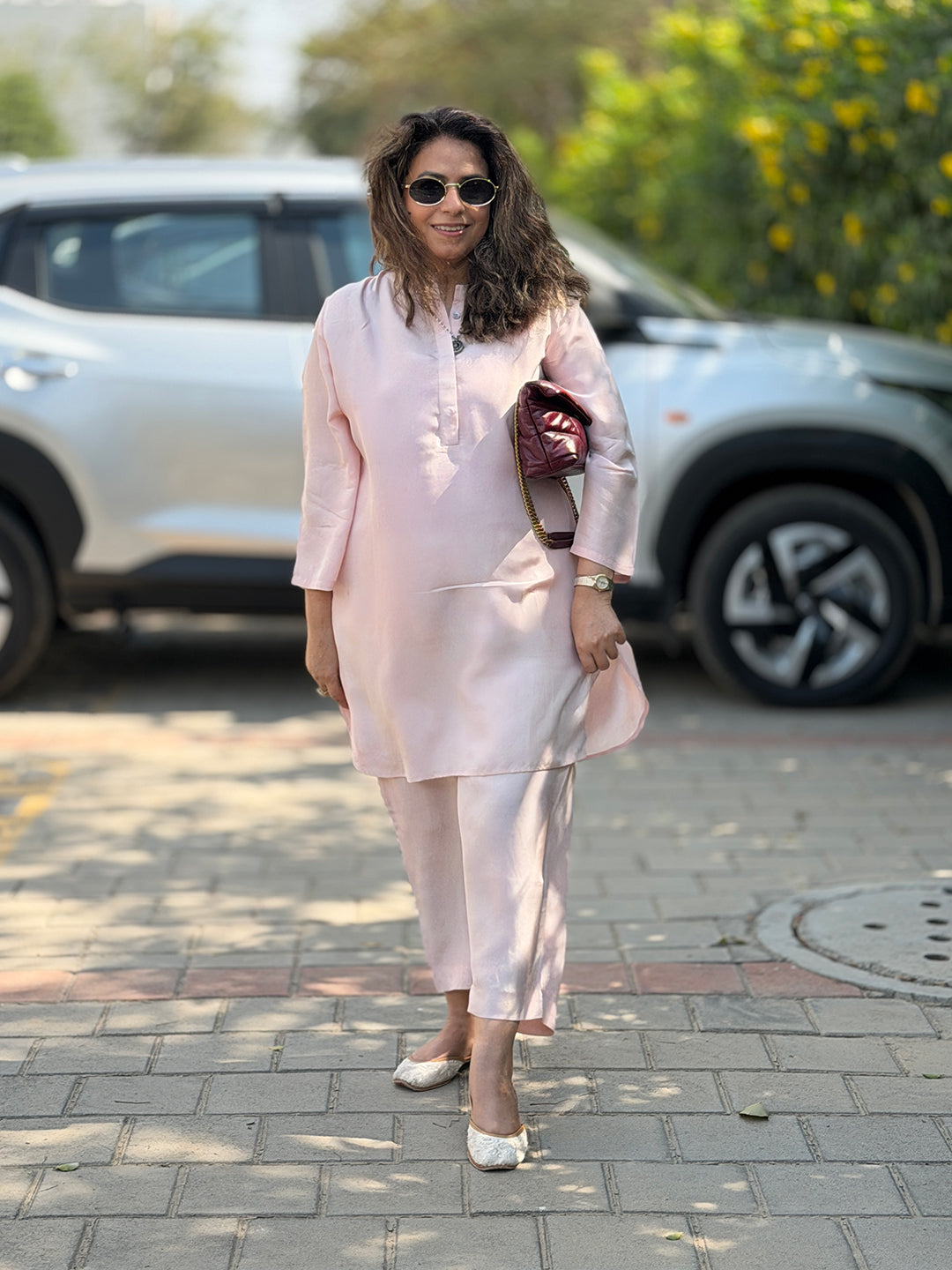 Woman in a light pink outfit standing on a paved area with a car and greenery in the background.