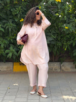 Woman in a light pink outfit standing outdoors with greenery in the background