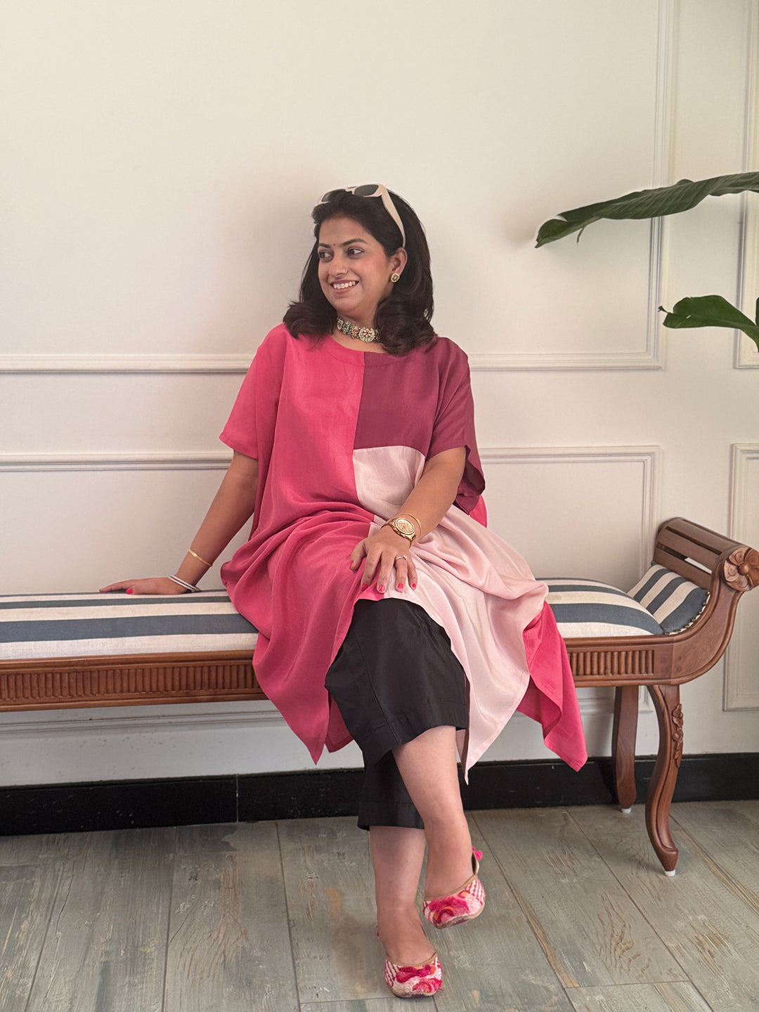 Woman sitting on a bench wearing a pink and black outfit with a white wall and plant in the background
