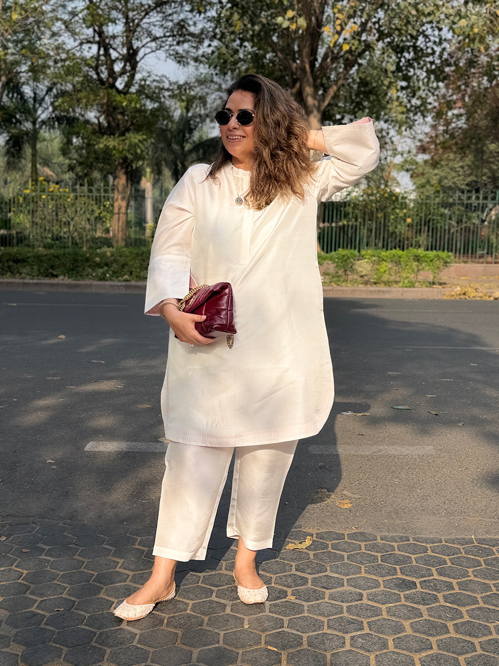 Woman in a white outfit holding a red clutch outdoors on a paved path with trees in the background.