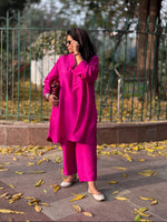 Woman in a bright pink traditional outfit standing on a pavement with leaves and a fence in the background.