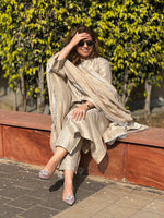 Woman in a beige traditional outfit sitting on a wall with greenery in the background
