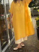 Woman in a yellow dress standing outdoors near a glass door and plants.