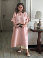 Woman in a pink dress holding a black Sabyasachi bag in a room with a wooden table and lamp.