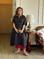 Woman in a black dress standing in a room with wooden flooring and a lamp in the background.
