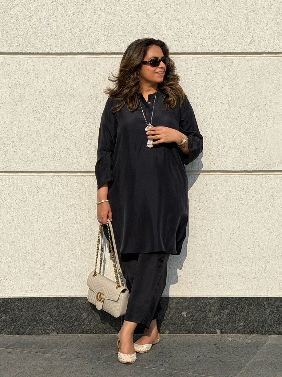 Woman in black outfit holding a beige handbag against a light-colored wall.