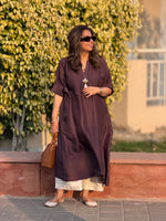 Woman in a long brown dress standing outdoors with greenery in the background