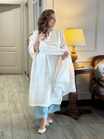 Woman in a white traditional outfit with blue pants standing in a room with wooden flooring and a lamp.