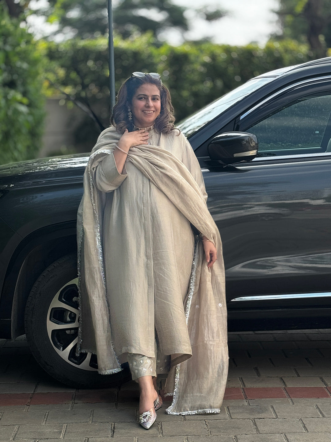 Woman in a beige traditional outfit standing next to a black car with greenery in the background