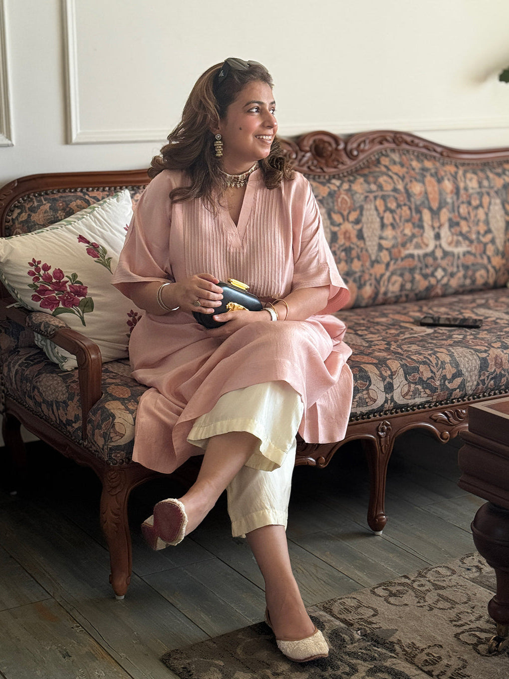 Woman sitting on a patterned sofa holding a Sabyasachi bag in a home setting.