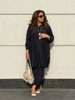 Woman in black outfit holding a beige handbag against a light-colored wall.