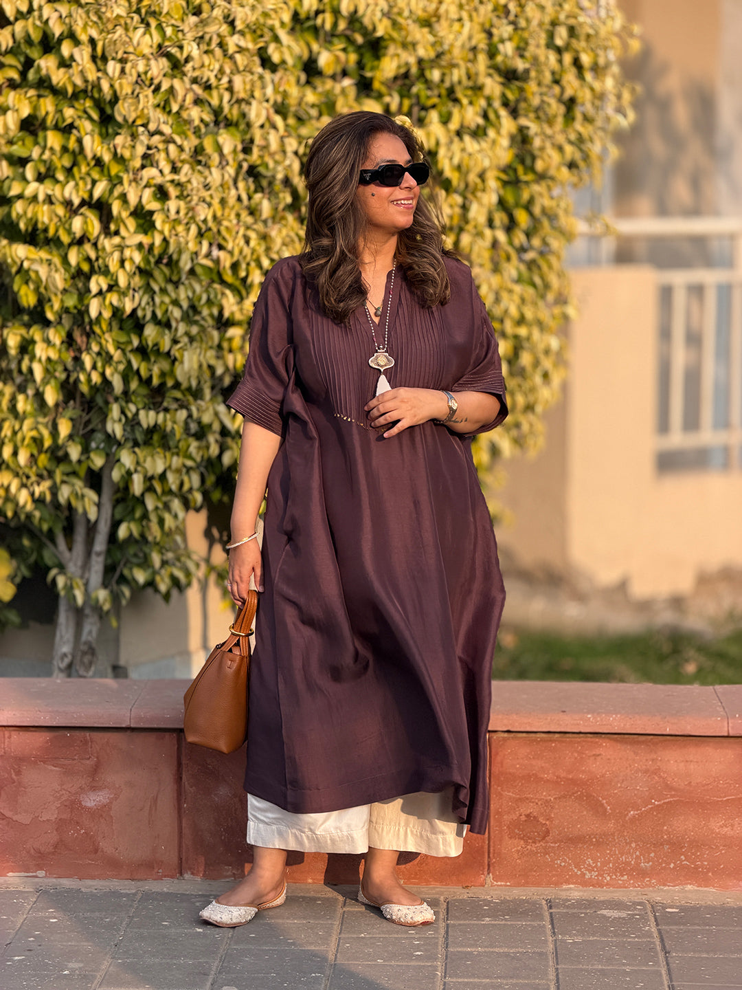 Woman in a long brown dress standing outdoors with greenery in the background