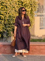 Woman in a long brown dress standing outdoors with greenery in the background