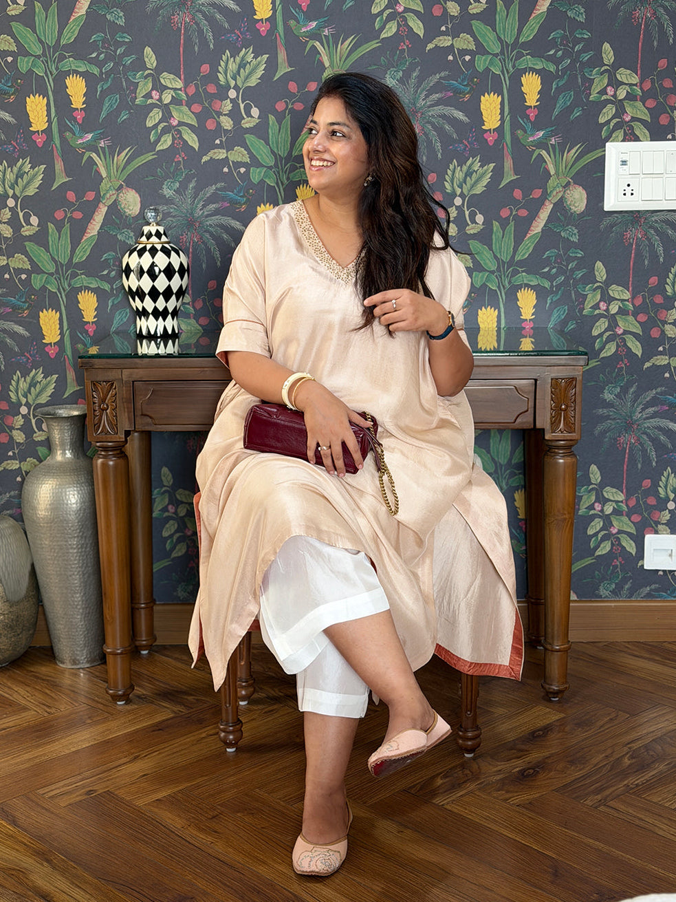 A woman sits on a wooden chair in front of a dark patterned wallpapered wall, wearing a light beige kurta with white cropped pants and holding a dark red clutch.