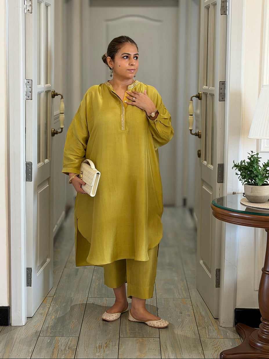 A woman wearing a mustard yellow kurta and matching trousers stands in a hallway framed by open white doors, holding a small cream clutch.