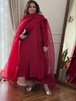 A woman stands in a room wearing a bright red traditional outfit with a matching sheer dupatta, holding a small black clutch. The setting features a wooden floor, patterned curtains, and a large mirror reflecting the outfit.