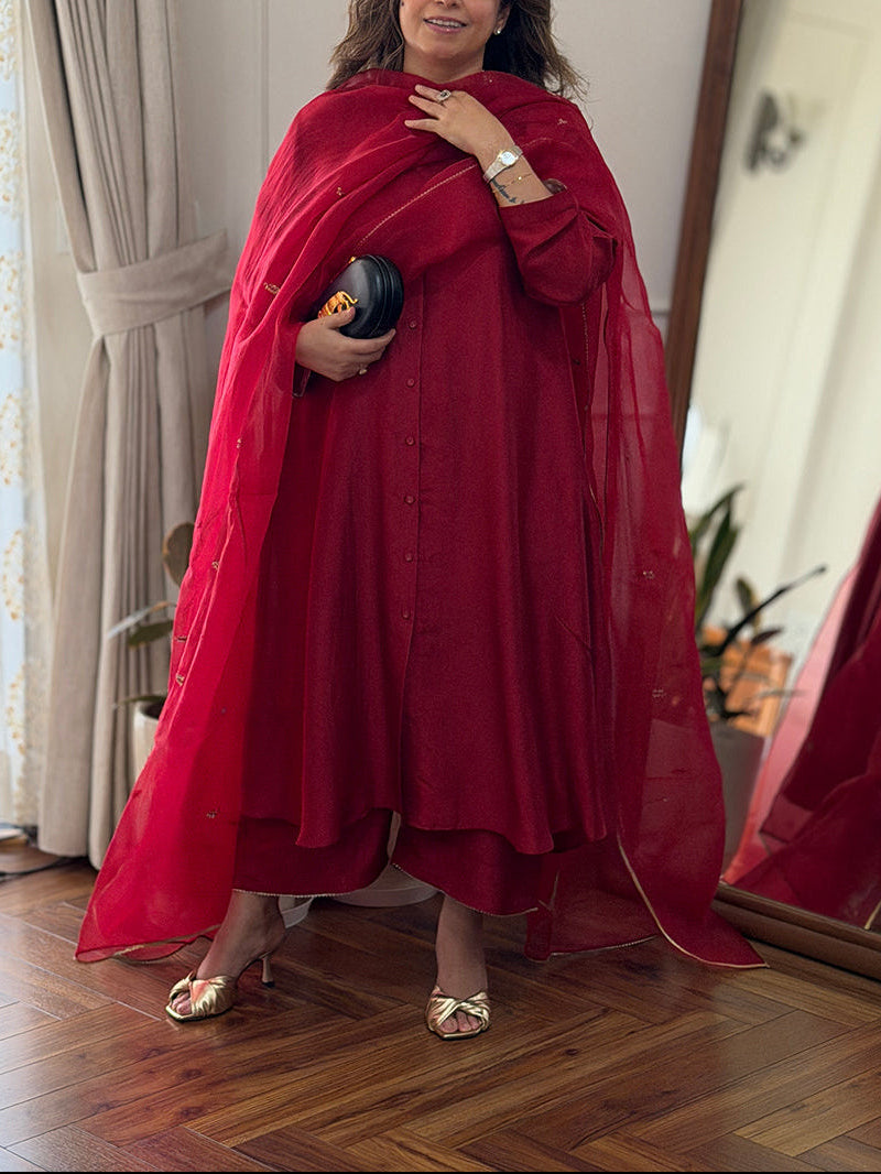 A woman wearing a vibrant red traditional outfit with a matching sheer dupatta and holding a black clutch, standing in a room with wooden floors, a large mirror, and curtains.