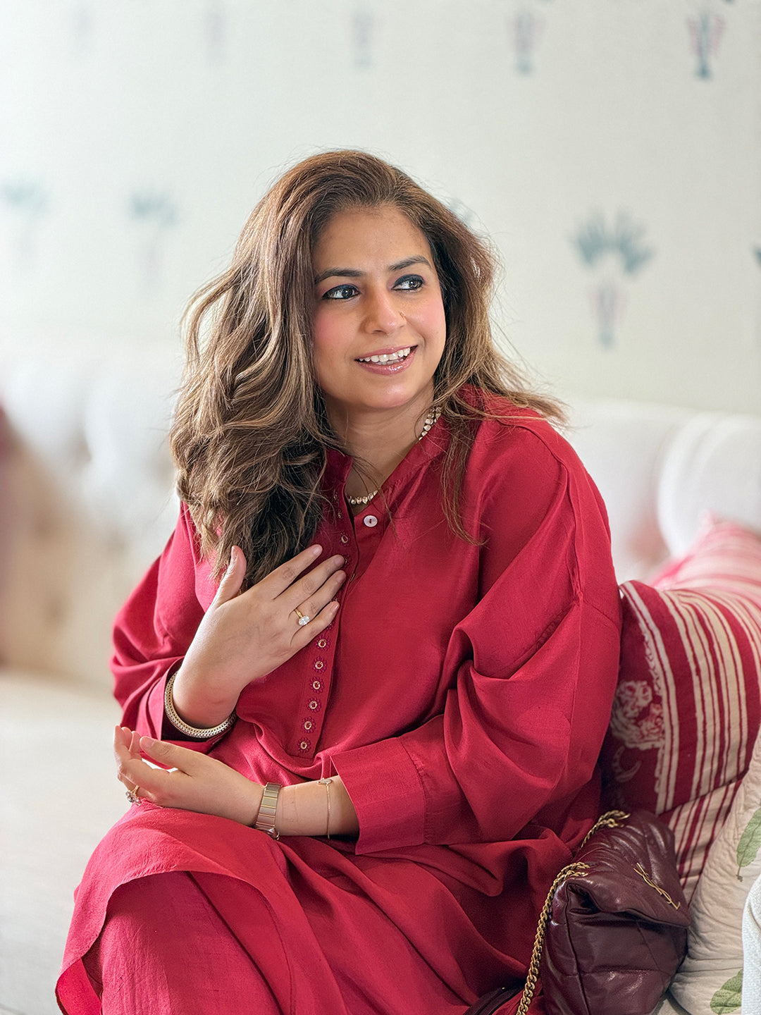 A woman with long wavy brown hair is seated on a sofa wearing a bright red long-sleeve tunic dress. She is smiling and looking to the side, with a neutral indoor background and a patterned cushion behind her.