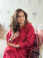 A woman with long wavy brown hair is seated on a sofa wearing a bright red long-sleeve tunic dress. She is smiling and looking to the side, with a neutral indoor background and a patterned cushion behind her.